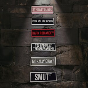 Puede incluir: Una colecci&oacute;n de letreros rectangulares con varias frases y palabras. Los letreros est&aacute;n montados en una pared de piedra texturizada de color gris oscuro. Las frases incluyen "STFUATTDLAGG", "I VOW. YOU VOW. WE VOW.", "DARK ROMANCE DR", "YOU HAD ME AT TRIGGER WARNING", "MORALLY GRAY ST" y "SMUT ST".