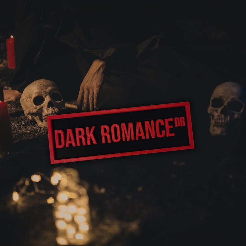 Puede incluir: Un letrero rectangular con las palabras "DARK ROMANCE" en letras rojas, enmarcado en rojo. Dos calaveras est&aacute;n a cada lado del letrero. La escena es oscura, con una mano visible.