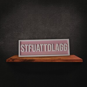 Puede incluir: Un letrero rectangular con el texto "STFUATTDLAGG" en letras blancas sobre un fondo rosa, enmarcado en blanco. El letrero descansa sobre una estanter&iacute;a de madera contra una pared gris oscuro.