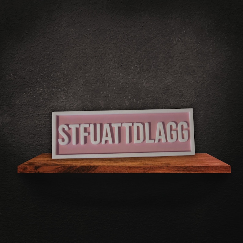 Puede incluir: Un letrero rectangular con el texto "STFUATTDLAGG" en letras blancas sobre un fondo rosa, enmarcado en blanco. El letrero descansa sobre una estanter&iacute;a de madera contra una pared gris oscuro.