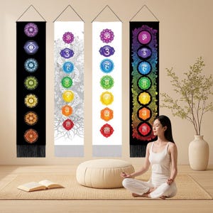 以下が含まれることがあります： カラフルなチャクラのシンボルとサンスクリット文字が描かれた、4つの垂直チャクラウォールハンギング。ハンギングは黒、白、グレーで、下部に黒いフリンジが付いています。女性がハンギングの前で瞑想しています。