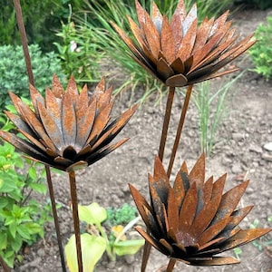 May include: Three metal garden stakes featuring stylized flower heads with pointed, layered petals. Each flower has a textured, rusted brown finish with dark gray undertones. The metal blooms are attached to thin, straight metal rods for support.
