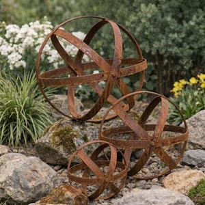 May include: Three rusty metal sphere garden ornaments of varying sizes. The spheres are constructed from intersecting bands, creating an open, orb-like structure. They are placed on a bed of rocks and gravel, with greenery in the background.