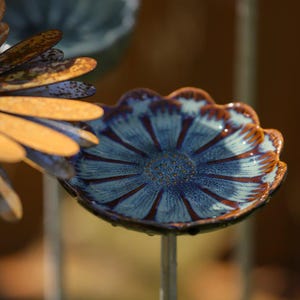 Puede incluir: Primer plano de un adorno de jardín con forma de flor de cerámica azul y marrón. La flor tiene un borde festoneado y un centro detallado. Los pétalos son azules con detalles marrones, montados en una varilla metálica.