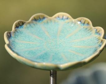 May include: A ceramic bird bath with a turquoise, flower-shaped dish. The dish has a scalloped edge with a yellow and blue trim. The bird bath is mounted on a metal rod, perfect for a garden or outdoor space.