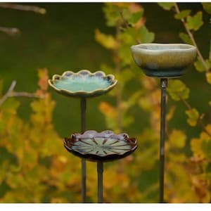 Könnte beinhalten: Drei Vogelbäder aus Keramik in verschiedenen Größen und Designs werden im Freien ausgestellt. Die Vogelbäder sind auf Metallstangen montiert und weisen eine Reihe von Farben auf, darunter Türkis, Braun und Gelb. Der Hintergrund ist mit grünem und gelbem Laub verschwommen.