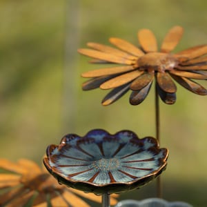 Può includere: Una scultura da giardino composta da un fiore in metallo con petali color marrone ruggine su uno stelo sottile, posizionato dietro un piatto in ceramica con bordo smerlato, smaltato in una finitura blu e marrone maculata con centro testurizzato.