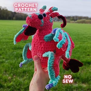 May include: A handmade crochet dragon in shades of red, teal, and purple. The dragon has large eyes and wings. The text "CROCHET PATTERN" and "NO SEW" are visible. The dragon is held in front of a green field.
