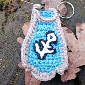 Peut inclure: Un porte-clés crocheté à la main avec un centre bleu clair orné d'un motif blanc et noir. Le porte-clés a une bordure beige et un porte-clés argenté. Le fond comprend des feuilles et une surface en bois.