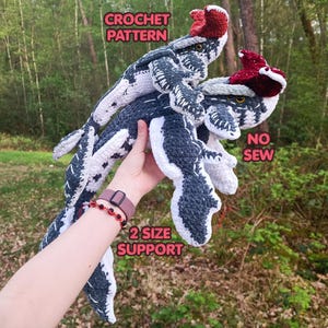 以下が含まれることがあります： 濃いグレーと白の糸で手編みされた2体のクロシェットの生き物で、赤いトサカが付いています。CROCHET PATTERN、NO SEW、2 SIZE SUPPORTという文字が見えます。生き物には詳細なヒレ、質感のある体、黄色い目があり、2つの異なるサイズで作られています。