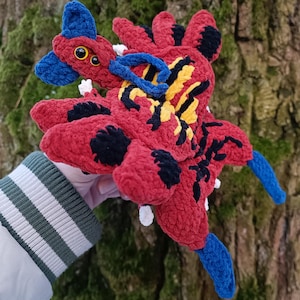 May include: A handmade plush creature in red, black, yellow, and blue. The plush has large black spots, yellow and black markings, and blue accents. It has two yellow eyes and is held in front of a tree.