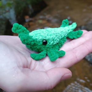 Puede incluir: Una criatura verde de ganchillo hecha a mano, con un cuerpo texturizado y ojos negros. El peluche es de un tono verde vibrante, con acentos verde oscuro. La criatura se sostiene en una mano, mostrando su tamaño y detalles.