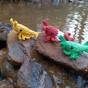 Puede incluir: Tres ajolotes de ganchillo en amarillo, rojo y verde descansan sobre una roca en un arroyo. Cada ajolote tiene una apariencia detallada y texturizada con pequeños ojos negros. El agua fluye suavemente alrededor de la roca.