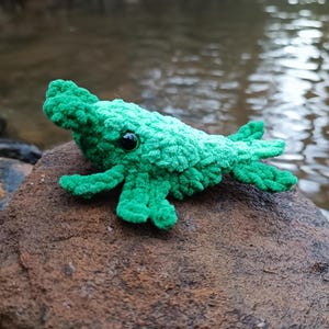 Puede incluir: Un juguete de cocodrilo verde tejido a mano con ojos de seguridad negros. El reptil de peluche está colocado sobre una roca marrón, con un fondo de agua borroso. El cocodrilo tiene un aspecto texturizado y difuso.