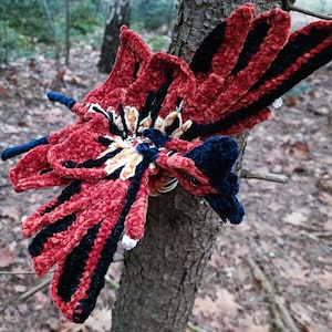 Könnte beinhalten: Ein handgefertigter, plüschiger Schmetterling in Rot-, Schwarz-, Weiß- und Blautönen, der auf einem Baumstamm sitzt. Der Schmetterling hat detaillierte Flügel und einen Körper mit samtiger Textur. Der Hintergrund zeigt eine verschwommene Waldszene.