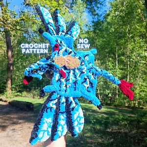 Könnte beinhalten: Ein blau-schwarzer Häkelvogel mit roten Akzenten. Der Vogel hat einen langen Schwanz und Flügel, mit dem Text "CROCHET PATTERN" und "NO SEW" sichtbar. Das Objekt ist aus weichem Garn gefertigt, was darauf hindeutet, dass es handgefertigt ist.