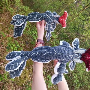 May include: Two handmade crochet creatures in a forest setting. The creatures are primarily dark blue with white and red accents. The crochet work features detailed stitching and unique shapes, resembling mythical beings.