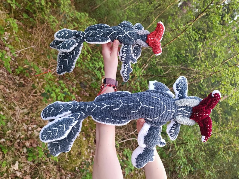 May include: Two handmade crochet creatures in a forest setting. The creatures are primarily dark blue with white and red accents. The crochet work features detailed stitching and unique shapes, resembling mythical beings.