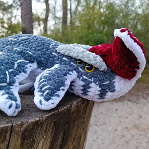 May include: A crocheted stuffed animal in the shape of a whale, featuring shades of blue, white, and gray. It has a red and white Santa hat and yellow eyes. The plush toy is resting on a wooden surface.