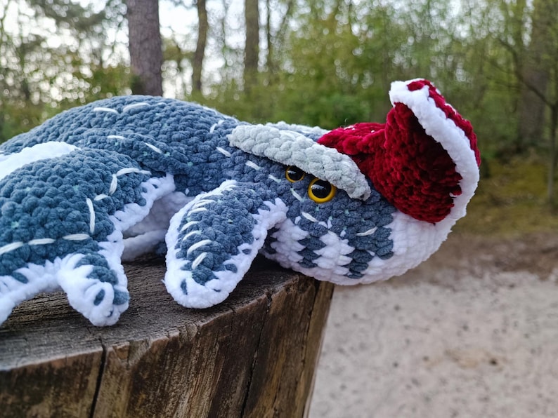 May include: A crocheted stuffed animal in the shape of a whale, featuring shades of blue, white, and gray. It has a red and white Santa hat and yellow eyes. The plush toy is resting on a wooden surface.