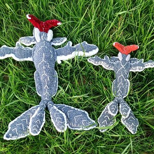 May include: Two handmade, gray and white crochet creatures with outstretched wings and red headpieces, resting on green grass. The creatures have a unique, whimsical design, perfect for a child's room or as a decorative item.