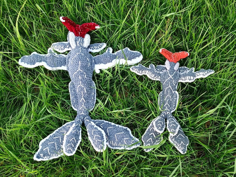 May include: Two handmade, gray and white crochet creatures with outstretched wings and red headpieces, resting on green grass. The creatures have a unique, whimsical design, perfect for a child's room or as a decorative item.