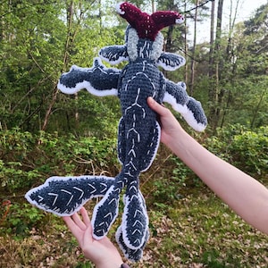 May include: A handmade crochet plush toy in the shape of a creature with a dark gray body, white accents, and a red headpiece. The toy has outstretched arms and a long tail, and is held against a backdrop of green foliage.