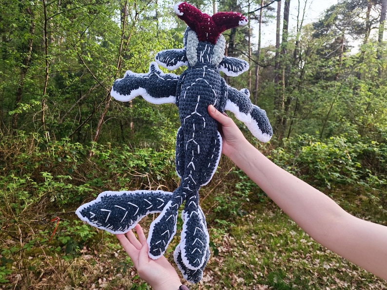 May include: A handmade crochet plush toy in the shape of a creature with a dark gray body, white accents, and a red headpiece. The toy has outstretched arms and a long tail, and is held against a backdrop of green foliage.