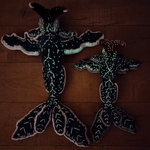 May include: Two plush, black creatures with outstretched arms and tail fins. The creatures have white dotted patterns and glowing accents. One has a red head, the other a black head. They are lying on a wooden surface.