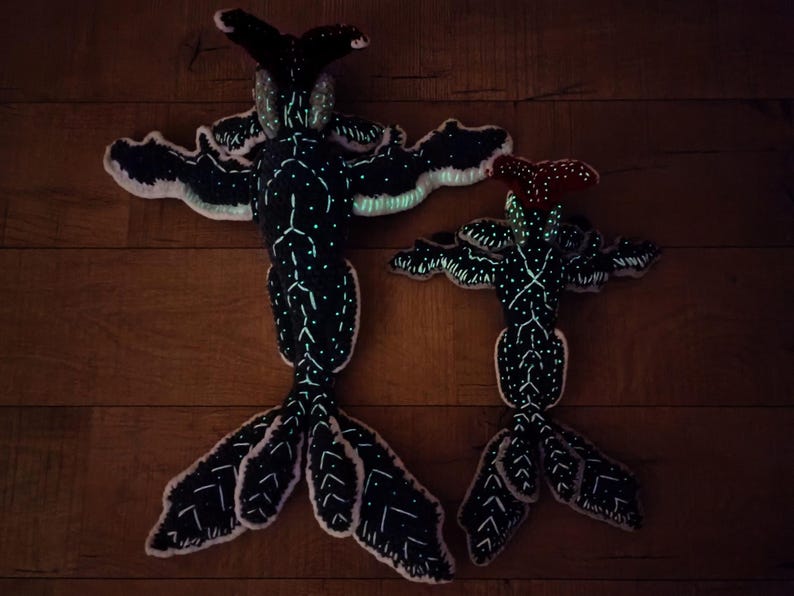 May include: Two plush, black creatures with outstretched arms and tail fins. The creatures have white dotted patterns and glowing accents. One has a red head, the other a black head. They are lying on a wooden surface.