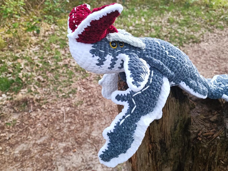 May include: A handmade crochet plush toy of a sea creature, primarily in shades of gray, white, and burgundy. The toy has a detailed design with white accents and yellow eyes. It is perched on a wooden surface, with a blurred natural background.