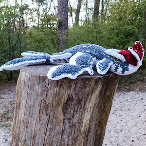 May include: A handmade crochet Nessie, the Loch Ness Monster, in shades of blue, gray, and white, wearing a red and white Santa hat. The plush toy is lying on a wooden tree stump, set against a backdrop of trees and foliage.