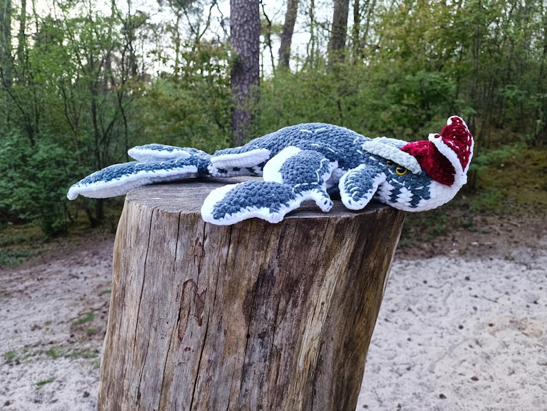 May include: A handmade crochet Nessie, the Loch Ness Monster, in shades of blue, gray, and white, wearing a red and white Santa hat. The plush toy is lying on a wooden tree stump, set against a backdrop of trees and foliage.