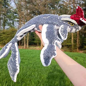 May include: A handmade, plush whale toy in shades of gray and white, with a red and white patterned headpiece. The whale is held up against a backdrop of green grass and trees, showcasing its soft, textured design. The toy features yellow eyes.