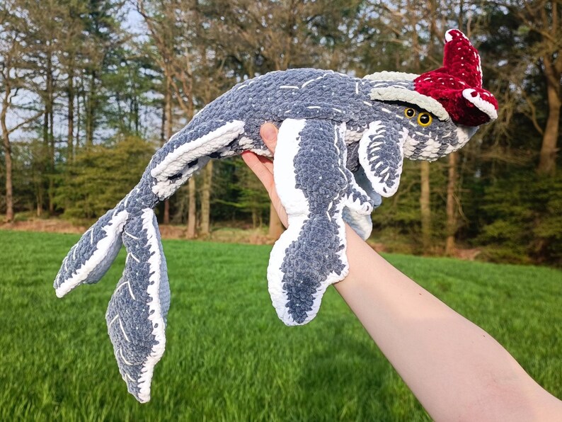 May include: A handmade, plush whale toy in shades of gray and white, with a red and white patterned headpiece. The whale is held up against a backdrop of green grass and trees, showcasing its soft, textured design. The toy features yellow eyes.