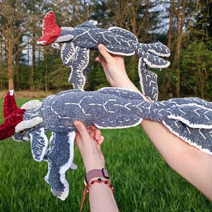 May include: Two handmade, plush, sea creature toys. The toys are primarily gray with white stitching and accents, and red details. The toys are held up against a backdrop of green grass and trees.