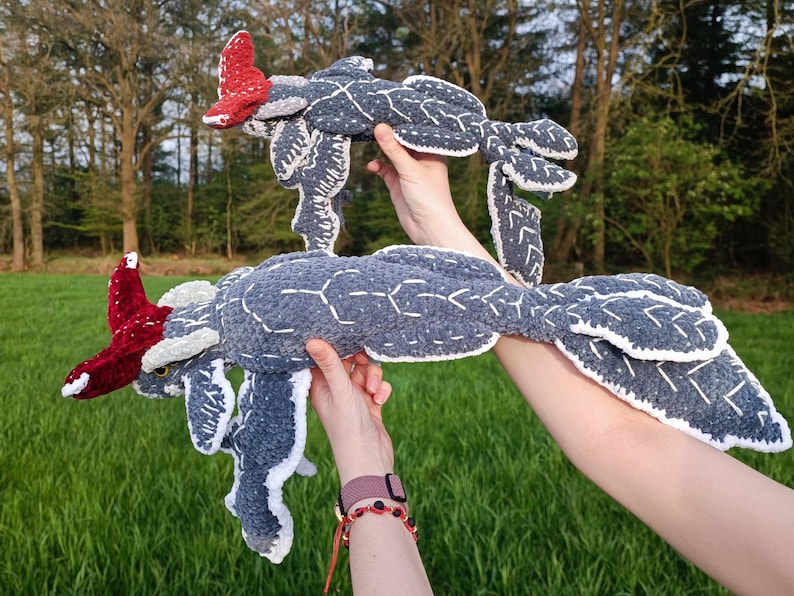 May include: Two handmade, plush, sea creature toys. The toys are primarily gray with white stitching and accents, and red details. The toys are held up against a backdrop of green grass and trees.