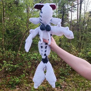 May include: A handmade crochet doll in a whimsical design. The doll features a white body with black spots, white arms and legs, and a dark blue tail. The head is white with a red and white accent. The doll is held up against a backdrop of green trees.