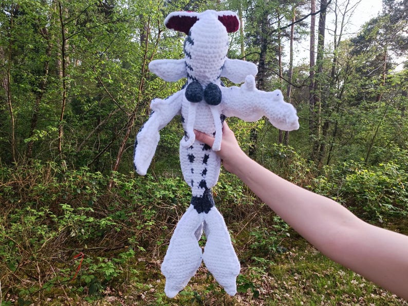 May include: A handmade crochet doll in a whimsical design. The doll features a white body with black spots, white arms and legs, and a dark blue tail. The head is white with a red and white accent. The doll is held up against a backdrop of green trees.
