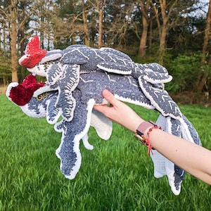 May include: A handmade crochet creature in shades of gray, white, and red. The plush toy has a detailed design with wings and a headpiece. The toy is held in front of a green grassy field and trees.