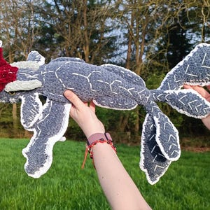 May include: A handmade, plush creature in shades of gray, white, and red. The body is a soft gray with white stitching, and the fins and tail have white edges. A red, textured headpiece adds a pop of color. The toy is held up against a backdrop of green grass and trees.