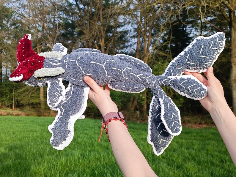 May include: A handmade, plush creature in shades of gray, white, and red. The body is a soft gray with white stitching, and the fins and tail have white edges. A red, textured headpiece adds a pop of color. The toy is held up against a backdrop of green grass and trees.