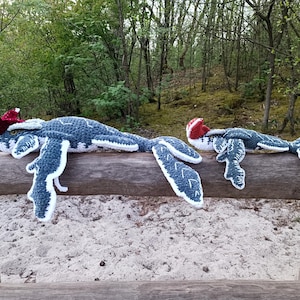 May include: Two handmade crochet sea serpent plush toys. The larger one is approximately 30 inches long, and the smaller one is about 20 inches long. They are blue and white with red accents on their heads, resting on a wooden fence.