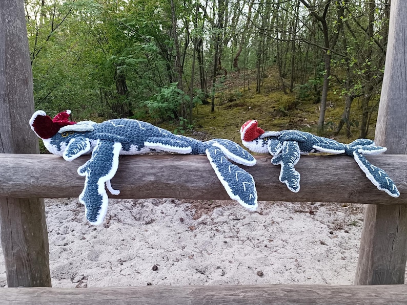 May include: Two handmade crochet sea serpent plush toys. The larger one is approximately 30 inches long, and the smaller one is about 20 inches long. They are blue and white with red accents on their heads, resting on a wooden fence.