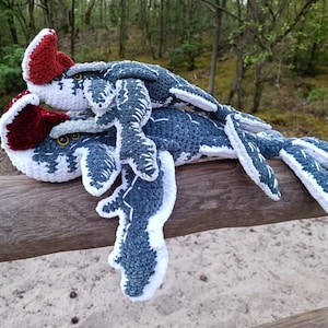 May include: Two crocheted plesiosaur plush toys in shades of blue, white, and burgundy rest on a wooden fence. The toys have detailed fins and heads with yellow eyes. The background features a blurred forest scene.