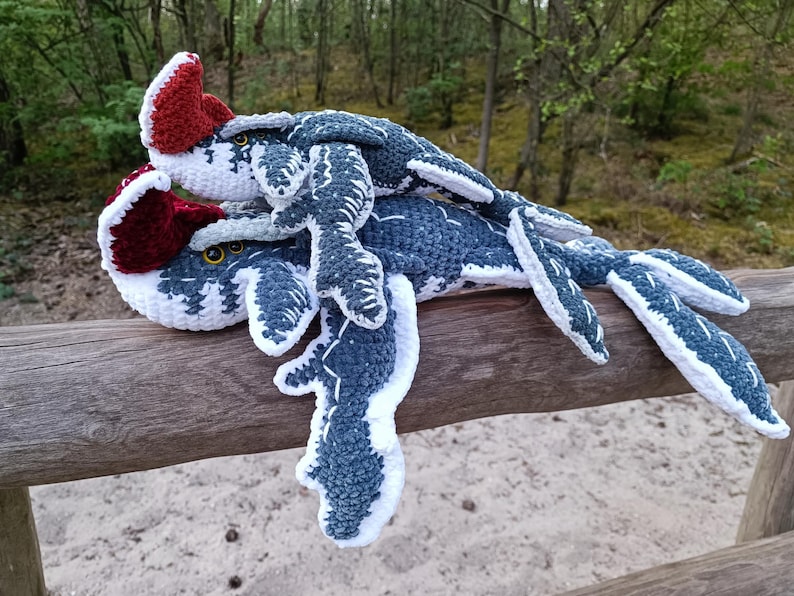 May include: Two crocheted plesiosaur plush toys in shades of blue, white, and burgundy rest on a wooden fence. The toys have detailed fins and heads with yellow eyes. The background features a blurred forest scene.