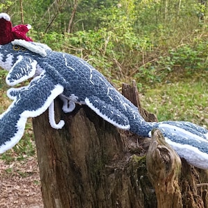 May include: A handmade crochet creature in shades of blue, white, and burgundy, resting on a tree stump. The whimsical design features a long body, wings, and a distinctive headpiece, showcasing intricate stitchwork and a playful aesthetic.