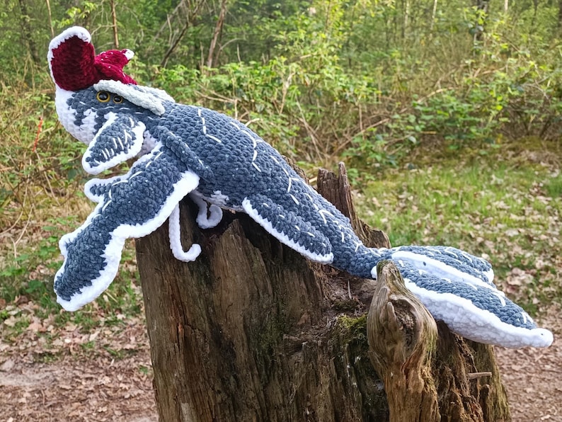 May include: A handmade crochet creature in shades of blue, white, and burgundy, resting on a tree stump. The whimsical design features a long body, wings, and a distinctive headpiece, showcasing intricate stitchwork and a playful aesthetic.