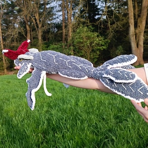 May include: A handmade, gray crocheted creature with white accents and a red headpiece. The plush toy has a geometric pattern and is held against a backdrop of green grass and trees.