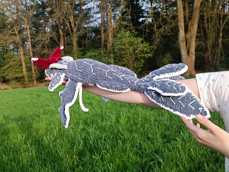 May include: A handmade, gray crocheted creature with white accents and a red headpiece. The plush toy has a geometric pattern and is held against a backdrop of green grass and trees.